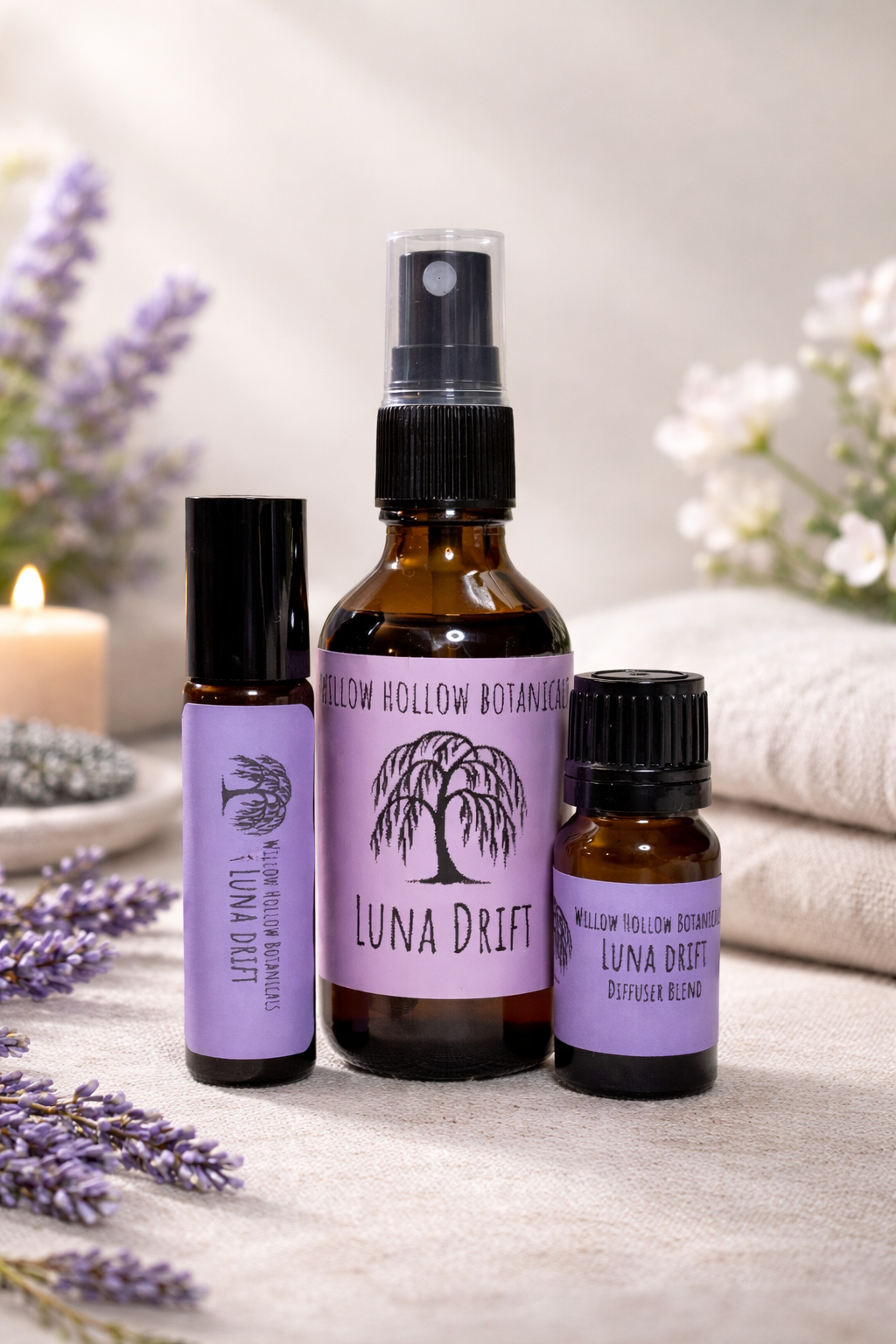 A studio photograph featuring three amber glass bottles from 'Willow Hollow Botanicals' with purple labels labeled 'Luna Drift.' The scent profile features a soothing, grounded blend of lavender, chamomile, and sandalwood. From left to right: a small rollerball bottle, a medium spray bottle, and a small dropper bottle labeled 'Diffuser Blend.' The bottles are arranged on a neutral fabric surface, surrounded by sprigs of fresh lavender and a soft-focus background featuring a glowing candle and white flowers.