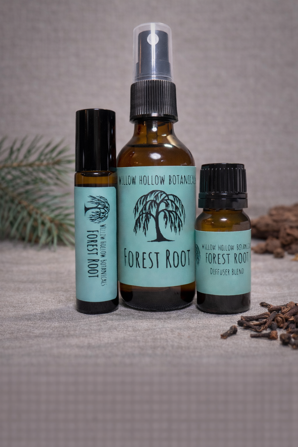 A studio photograph featuring three amber glass bottles from 'Willow Hollow Botanicals' with seafoam green labels labeled 'Forest Root.' The scent profile is a crisp, woodsy blend of refreshing pine, grounding cedarwood, and warm, spiced clove. From left to right: a small rollerball bottle, a medium spray bottle, and a small dropper bottle labeled 'Diffuser Blend.' The bottles are arranged on a neutral surface, with a pine sprig on the left and scattered whole cloves on the right.