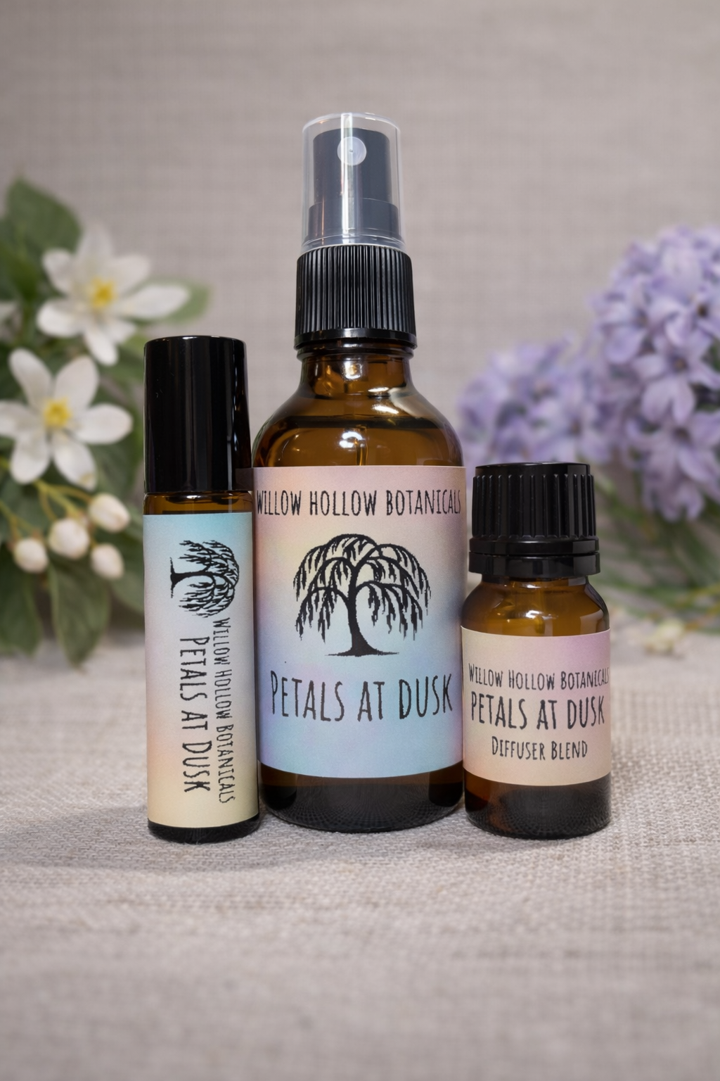 A studio photograph featuring three amber glass bottles from 'Willow Hollow Botanicals' with iridescent, sunset-hued labels labeled 'Petals at Dusk.' The scent profile is a floral blend featuring jasmine, hyacinth, and rose, grounded by amber and oak moss. From left to right: a small rollerball bottle, a medium spray bottle, and a small dropper bottle labeled 'Diffuser Blend.' The bottles are arranged on a neutral fabric surface, framed by white and purple floral accents in the soft-focus background.