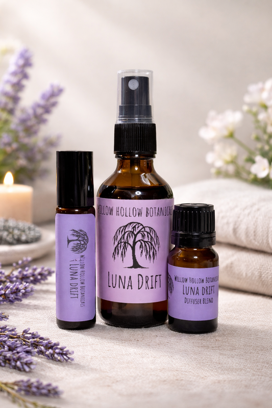 A studio photograph featuring three amber glass bottles from 'Willow Hollow Botanicals' with purple labels labeled 'Luna Drift.' The scent profile features a soothing, grounded blend of lavender, chamomile, and sandalwood. From left to right: a small rollerball bottle, a medium spray bottle, and a small dropper bottle labeled 'Diffuser Blend.' The bottles are arranged on a neutral fabric surface, surrounded by sprigs of fresh lavender and a soft-focus background featuring a glowing candle and white flowers.