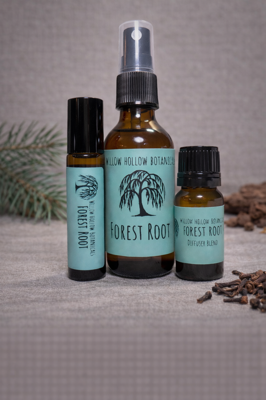 A studio photograph featuring three amber glass bottles from 'Willow Hollow Botanicals' with seafoam green labels labeled 'Forest Root.' The scent profile is a crisp, woodsy blend of refreshing pine, grounding cedarwood, and warm, spiced clove. From left to right: a small rollerball bottle, a medium spray bottle, and a small dropper bottle labeled 'Diffuser Blend.' The bottles are arranged on a neutral surface, with a pine sprig on the left and scattered whole cloves on the right.
