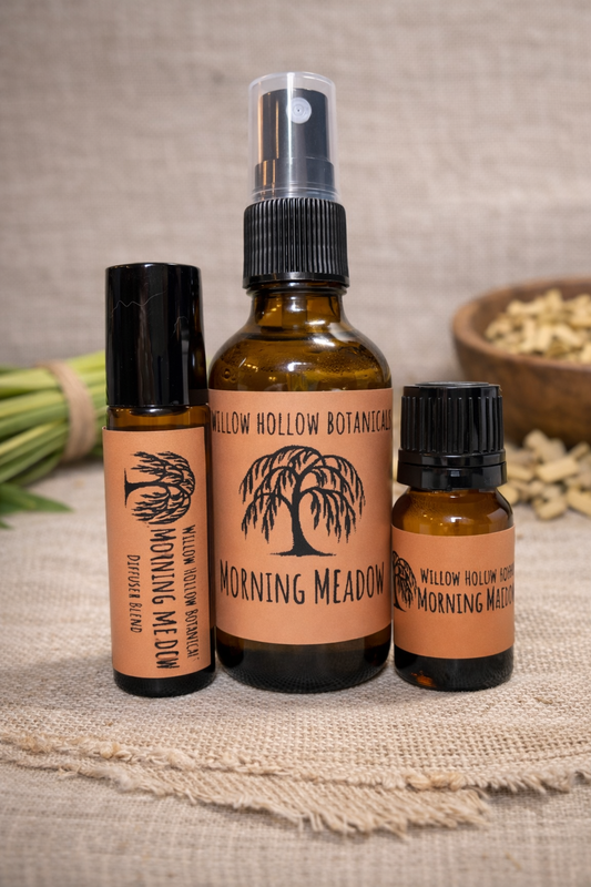 A studio photograph featuring three amber glass bottles from 'Willow Hollow Botanicals' with peach-toned labels labeled 'Morning Meadow.' The scent profile features the fresh, zesty, and revitalizing aroma of pure lemongrass. From left to right: a small rollerball bottle, a medium spray bottle, and a small dropper bottle labeled 'Diffuser Blend.' The bottles are arranged on a rustic burlap surface, with a bundle of fresh lemongrass stalks on the left and a small wooden bowl of dried herbs in the background.