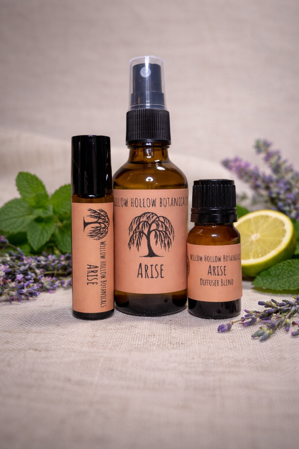 A studio photograph featuring three amber glass bottles from 'Willow Hollow Botanicals' with peach-toned labels labeled 'Arise.' The scent profile is a fresh, uplifting blend of cooling spearmint, calming lavender, and bright, citrusy bergamot. From left to right: a small rollerball bottle, a medium spray bottle, and a small dropper bottle labeled 'Diffuser Blend.' The bottles are arranged on a neutral fabric surface, surrounded by fresh mint leaves, sprigs of lavender, and a slice of lime in the background