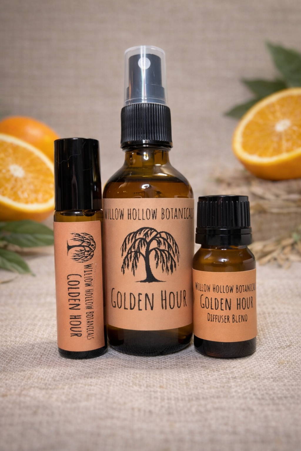 A studio photograph featuring three amber glass bottles from 'Willow Hollow Botanicals' with peach-toned labels labeled 'Golden Hour.' The scent profile features the bright, sunny, and joyful aroma of sweet orange. From left to right: a small rollerball bottle, a medium spray bottle, and a small dropper bottle labeled 'Diffuser Blend.' The bottles are arranged on a textured burlap surface, framed by fresh slices of oranges in the background to emphasize the citrus notes.
