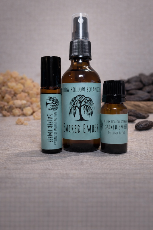 A studio photograph featuring three amber glass bottles from 'Willow Hollow Botanicals' with seafoam green labels labeled 'Sacred Ember.' The scent profile is a warm, mystical blend of resinous frankincense, earthy myrrh, and sweet, spiced tonka bean. From left to right: a small rollerball bottle, a medium spray bottle, and a small dropper bottle labeled 'Diffuser Blend.' The bottles are arranged on a neutral surface, flanked by piles of frankincense resin on the left and dried tonka beans on the right.