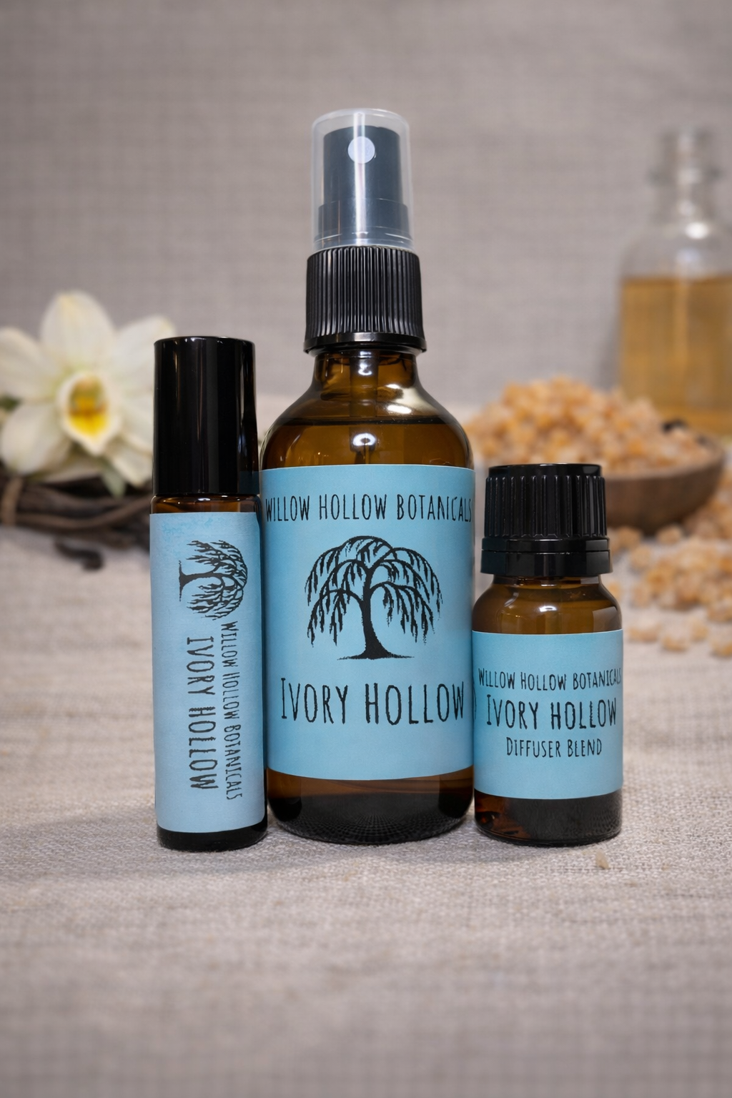 A studio photograph featuring three amber glass bottles from 'Willow Hollow Botanicals' with light blue labels labeled 'Ivory Hollow.' The scent profile is a cozy, crisp blend of fresh black linen, warm amber, and sweet vanilla. From left to right: a small rollerball bottle, a medium spray bottle, and a small dropper bottle labeled 'Diffuser Blend.' The bottles are arranged on a rustic, textured fabric surface, with a delicate white flower and a small bowl of aromatic resin in the soft-focus background.