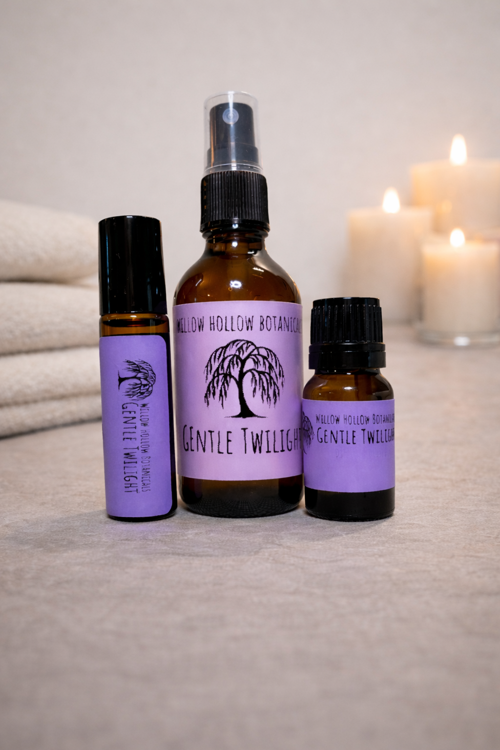 A studio photograph featuring three amber glass bottles from 'Willow Hollow Botanicals' with purple labels labeled 'Gentle Twilight.' The scent profile features a deep, calming blend of vanilla, frankincense, jasmine, and  tonka bean. From left to right: a small rollerball bottle, a medium spray bottle, and a small dropper bottle. The bottles are arranged on a neutral surface with folded white towels on the left and three glowing, out-of-focus candles in the background, creating a calm, spa-like atmosphere.