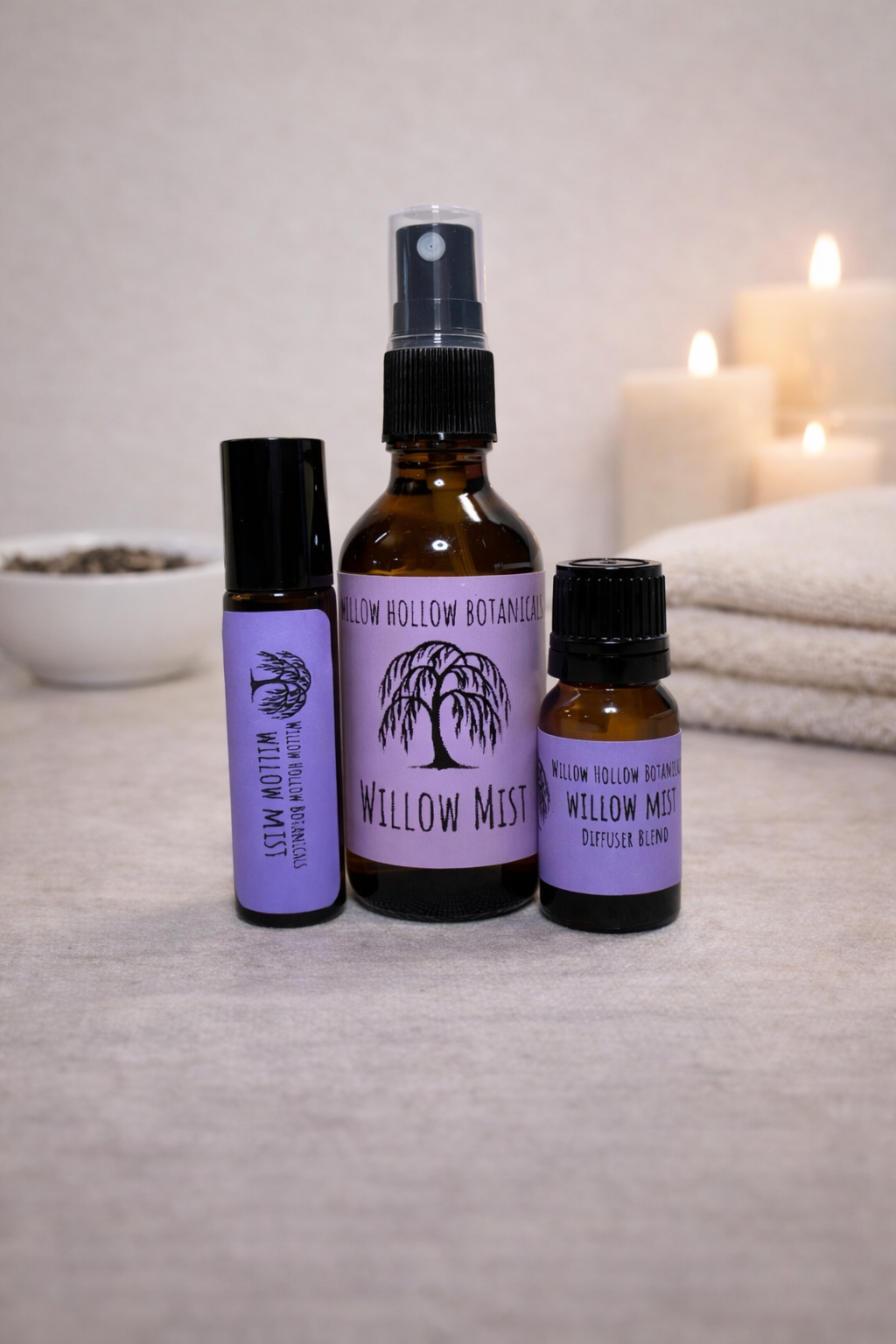 A studio photograph featuring three amber glass bottles from 'Willow Hollow Botanicals' with purple labels labeled 'Willow Mist.' The blend features soothing notes of white tea, bright bergamot, and warm, sweet vanilla. From left to right: a small rollerball bottle, a medium spray bottle, and a small dropper bottle labeled 'Diffuser Blend.' The bottles are arranged on a neutral surface with a small white bowl of dried botanicals on the left and folded white towels and glowing candles in the background.
