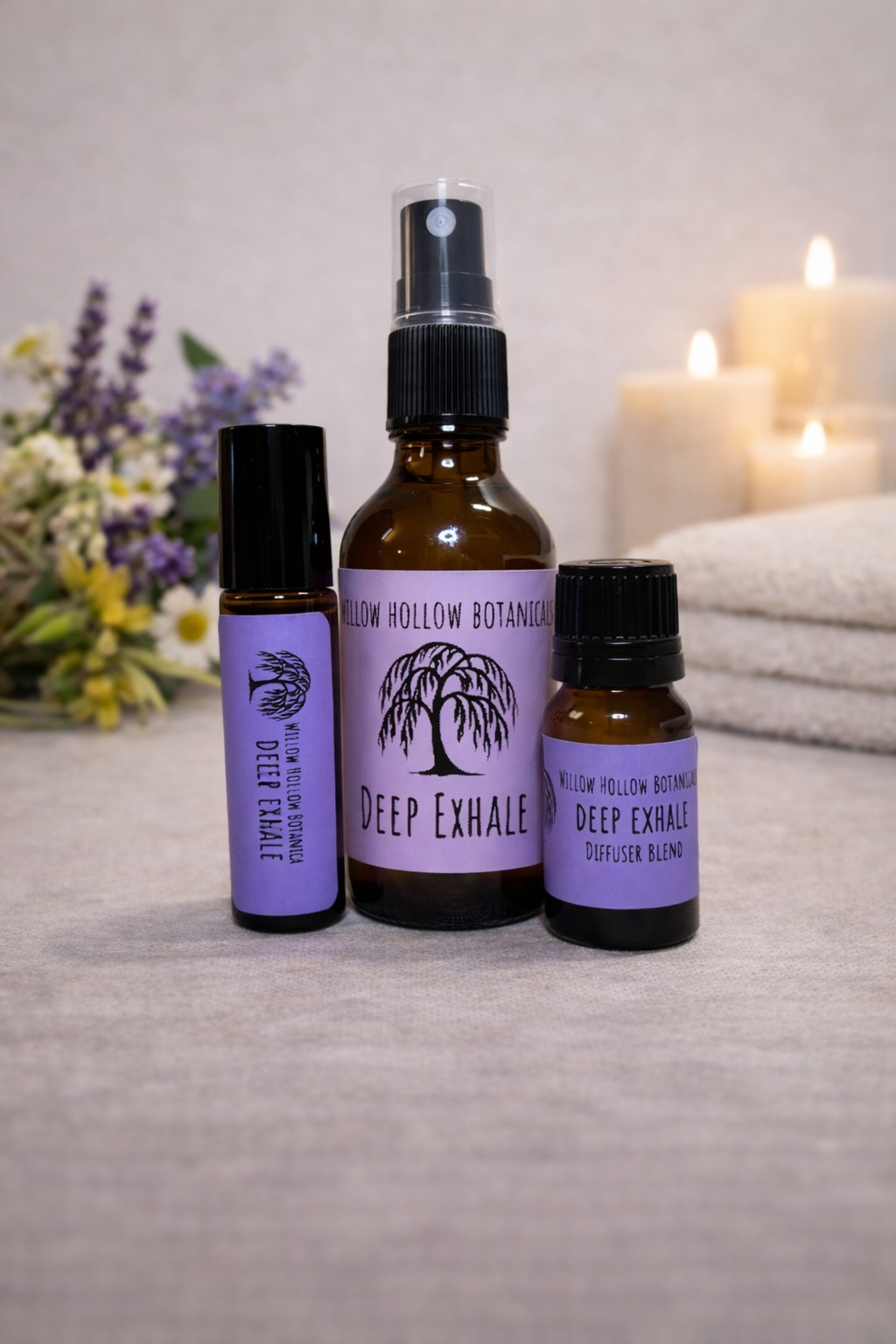 A studio photograph featuring three amber glass bottles from 'Willow Hollow Botanicals' with purple labels labeled 'Deep Exhale.' This blend features a grounding profile of lavender, sage, vetiver, chamomile, and bergamot. From left to right: a small rollerball bottle, a medium spray bottle, and a small dropper bottle labeled 'Diffuser Blend.' The bottles are arranged on a neutral surface next to a small bouquet of fresh wildflowers, with soft-focus folded white towels and glowing candles in the background.