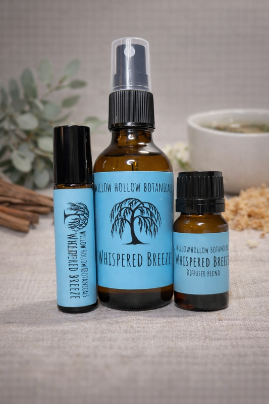 A studio photograph featuring three amber glass bottles from 'Willow Hollow Botanicals' with light blue labels labeled 'Whispered Breeze.' The scent profile is a light, calming blend of white tea, eucalyptus, and vanilla. From left to right: a small rollerball bottle, a medium spray bottle, and a small dropper bottle labeled 'Diffuser Blend.' The bottles are arranged on a rustic, textured fabric surface, with sprigs of greenery and a small white ceramic bowl in the soft-focus background.